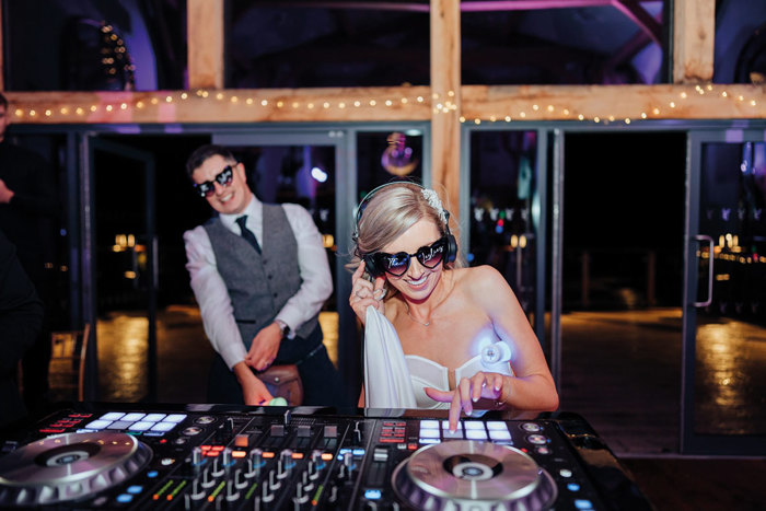 Bride on DJ decks and groom watching, both wearing sunglasses
