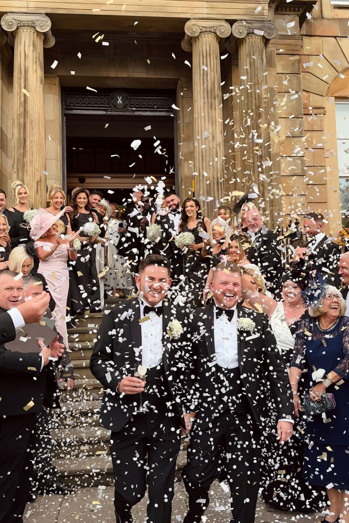 two grooms smile as wedding guests throw confetti over them in glasgow city centre