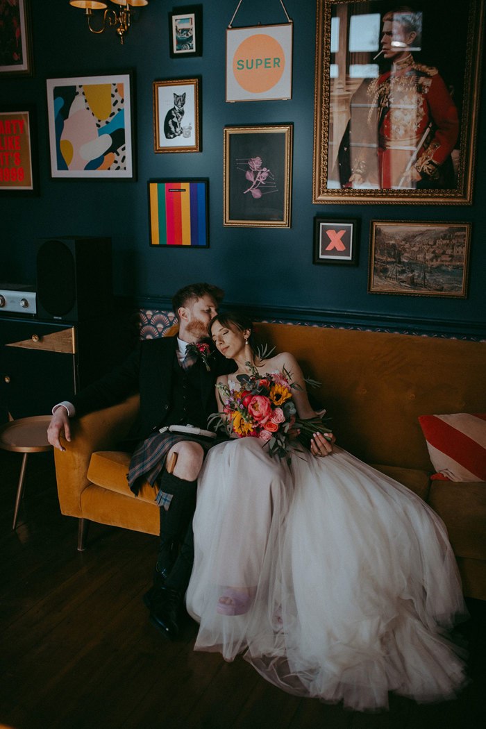 a bride and groom sitting on a yellow velvet sofa below a wall filled with colourful modern artwork at Netherbyres House