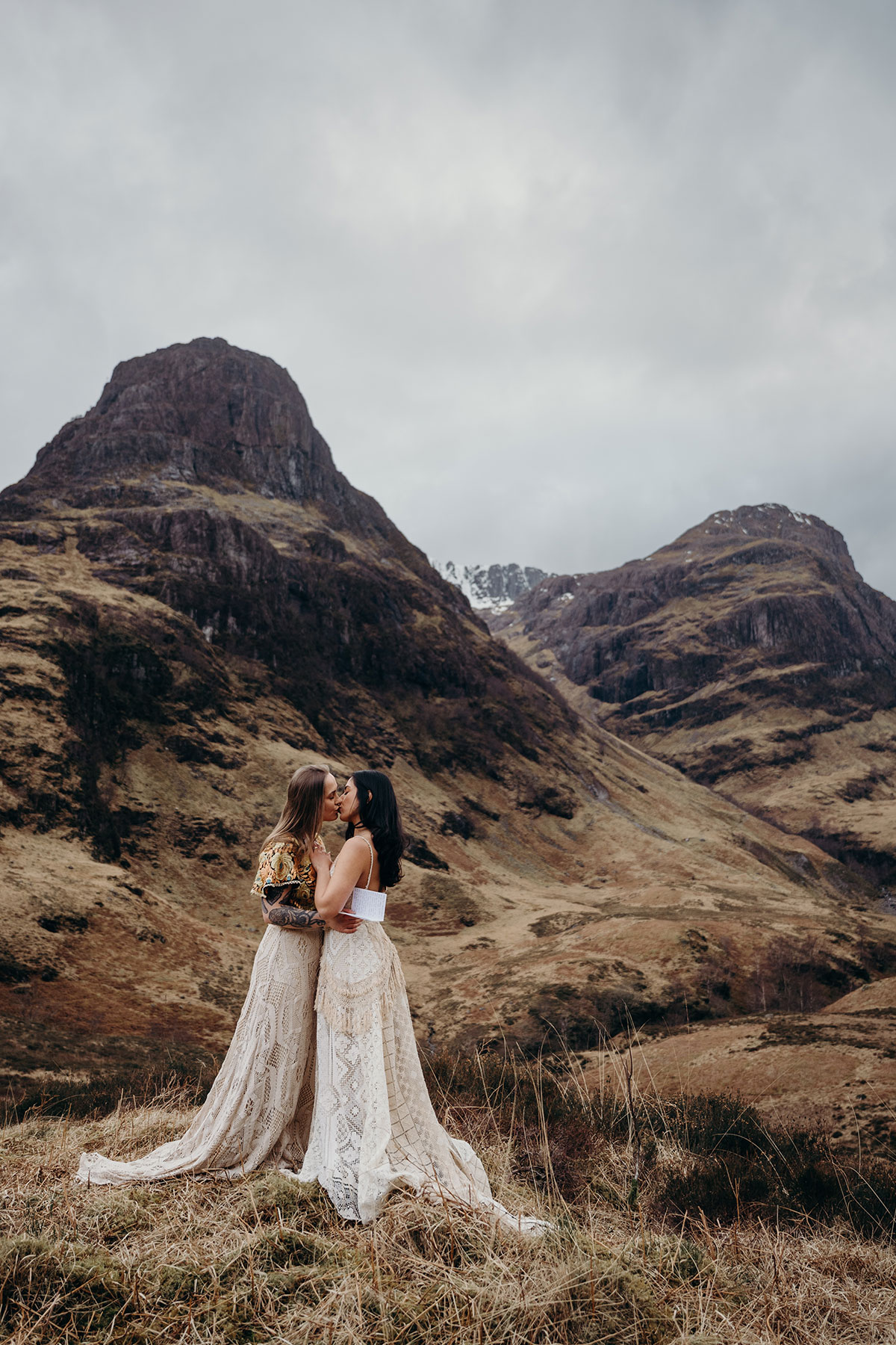 Couple kissing in lace wedding outfits during a Glencoe elopement shoot with dramatic Scottish mountain scenery behind them.