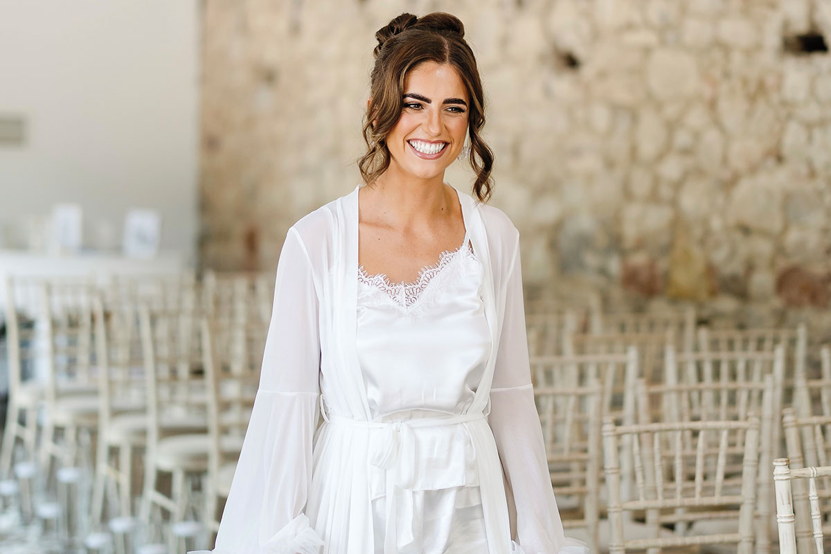 Bride smiling in a white satin camisole and sheer robe while getting ready in a light-filled ceremony space