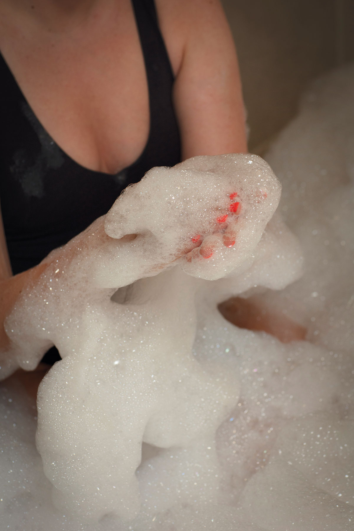 woman in black low neck swimsuit with neon pink fingernails sits immersed in a room full of soapy bubbles
