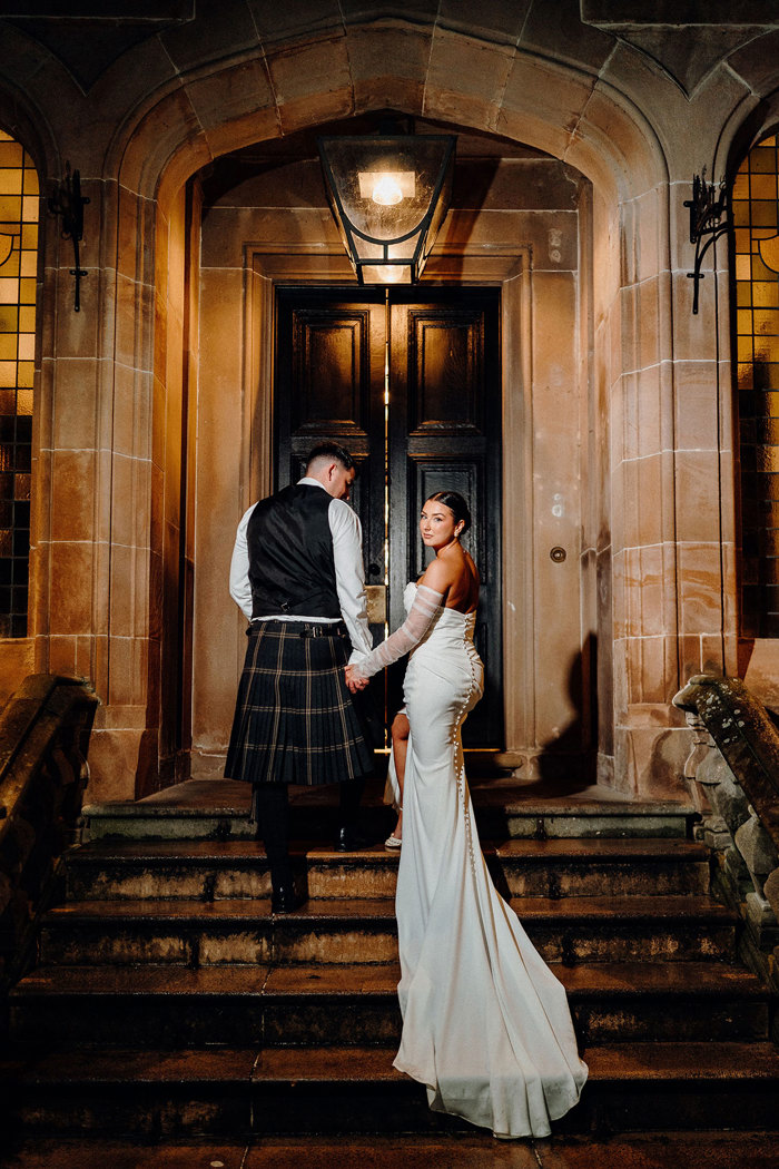 bride in strapless wedding dress with detached tulle sleeves looks back at camera while standing on outdoor set of stairs beside tall groom in beige and dark green kilt outfit