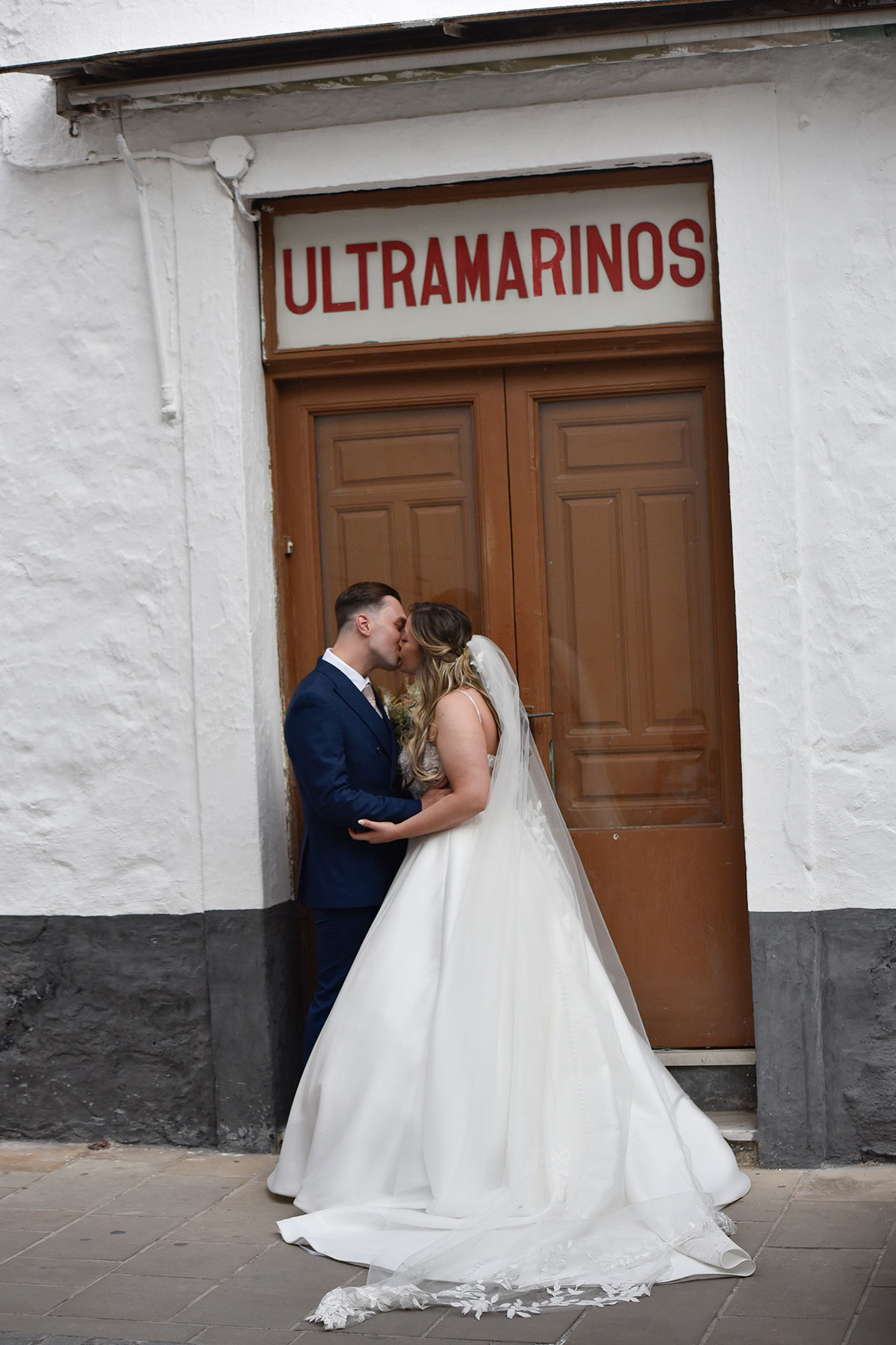 bride in white ballgown stands and trailing floral applique veil kisses groom in blue suit stood outside wooden doors that say 'ultramarinos' on a sign above the doorway