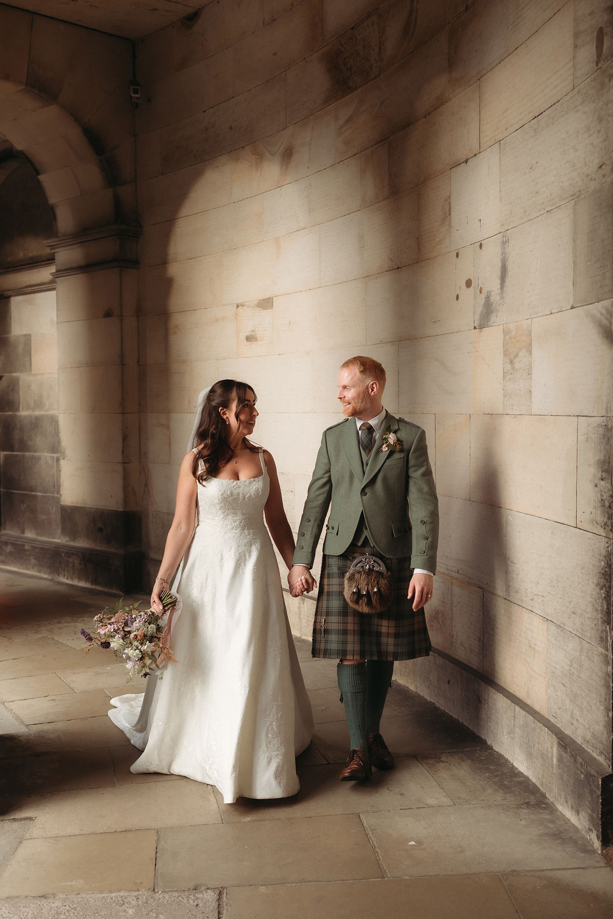 a bride and groom walking hand in hand outside a pale stone building