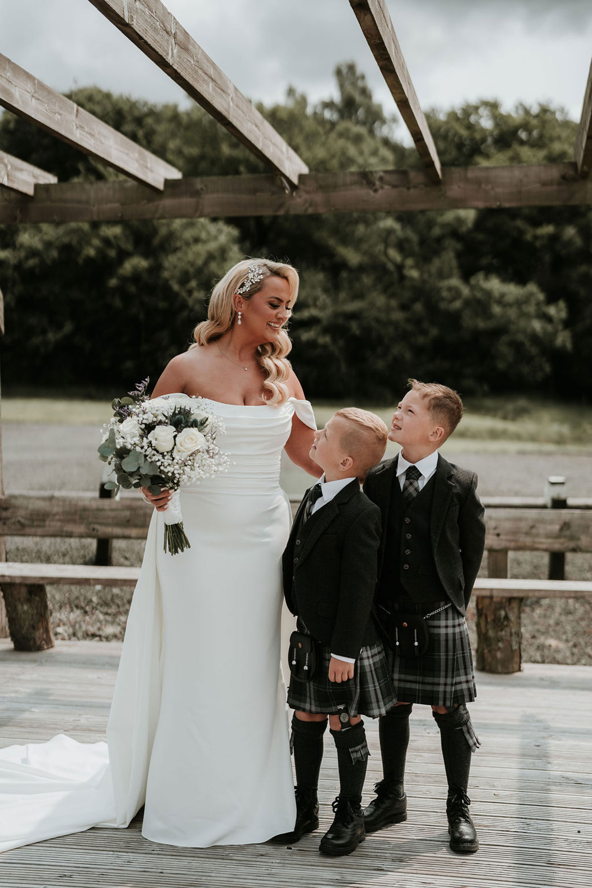 Bride standing with two young boys in kilts during outdoor wedding portraits.