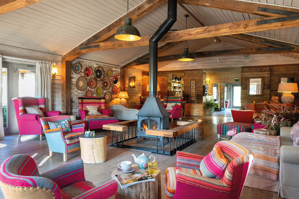 colourful wooden cottage with wood burning stove in centre with bright pink and striped lounge furniture scattered around the room
