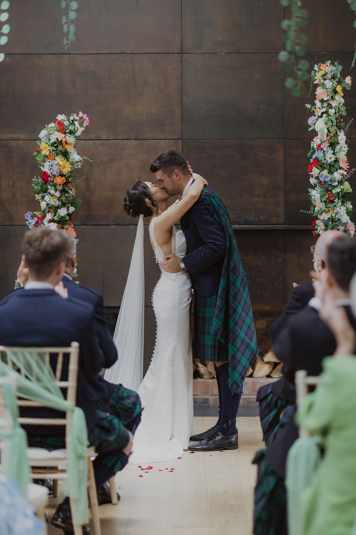 Newlyweds share their first kiss at indoor wedding ceremony with guests seated and floral pillars framing the aisle.
