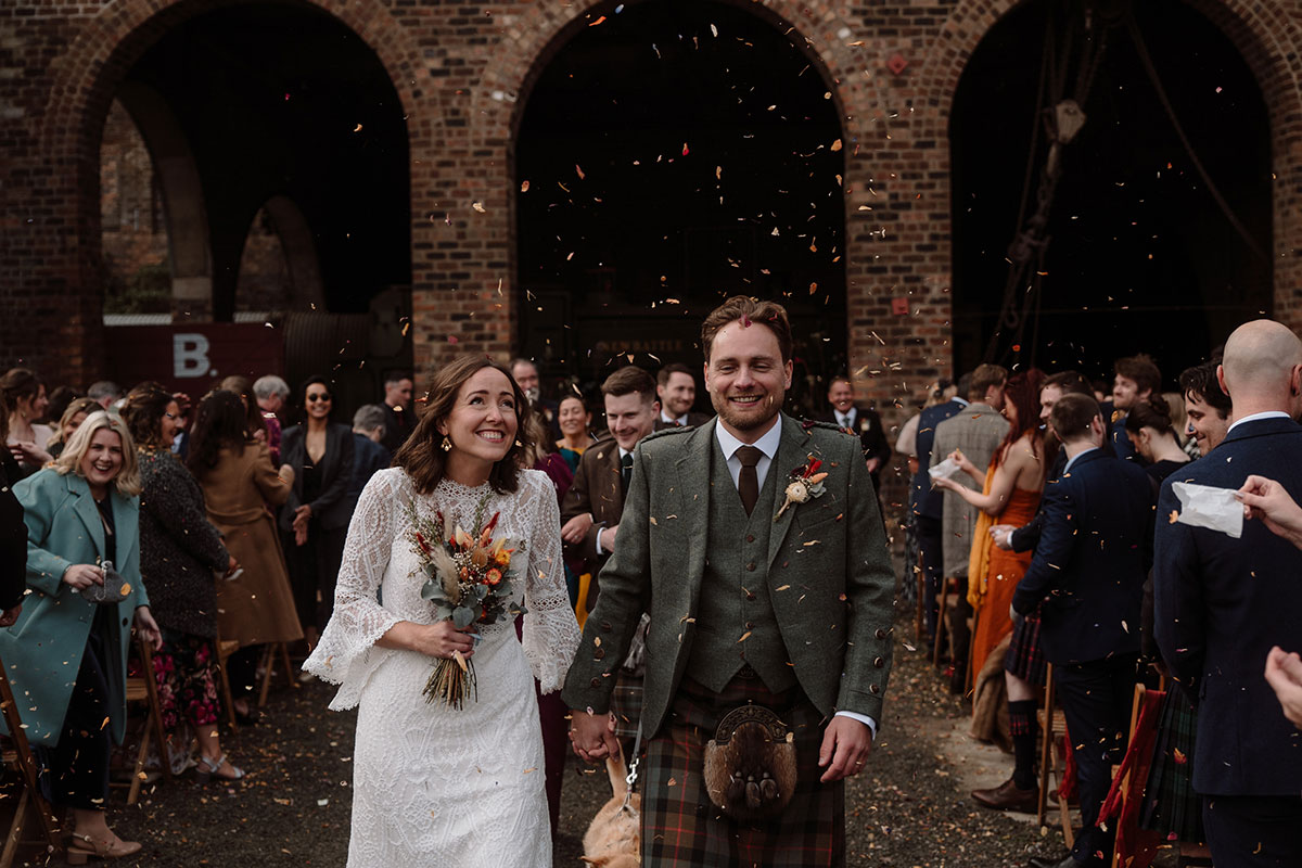 Newlyweds walk through a confetti shower outside an industrial wedding venue, surrounded by family and friends after their wedding ceremony