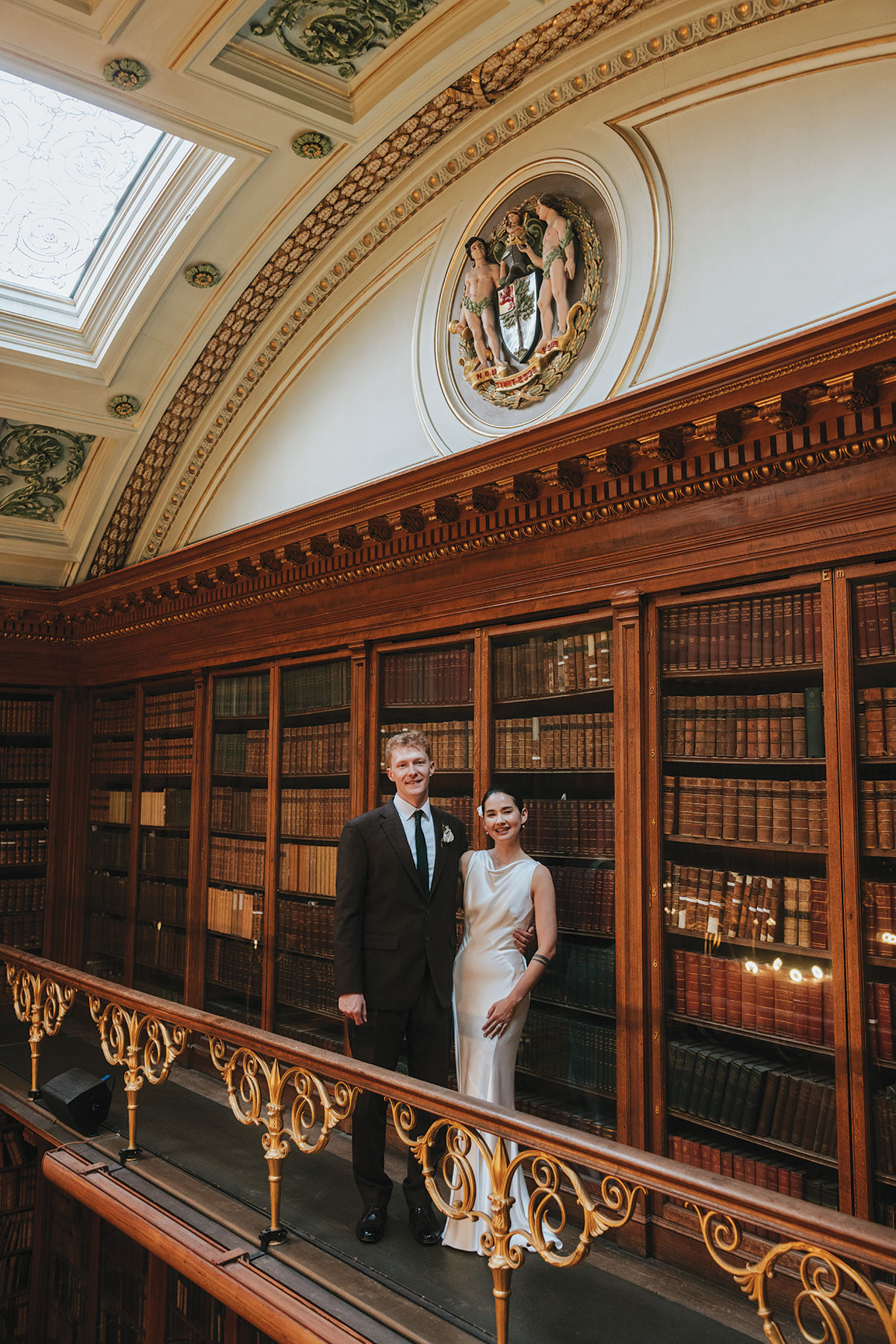 Bride and groom standing together on a balcony in a historic library with tall bookshelves.