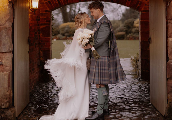 blonde bride in feathery cape and white wedding dress holds bouquet of white roses and stands smiling and facing groom in green jacket and red, blue, green kilt with matching fly plaid, both stood under outdoor stone archway