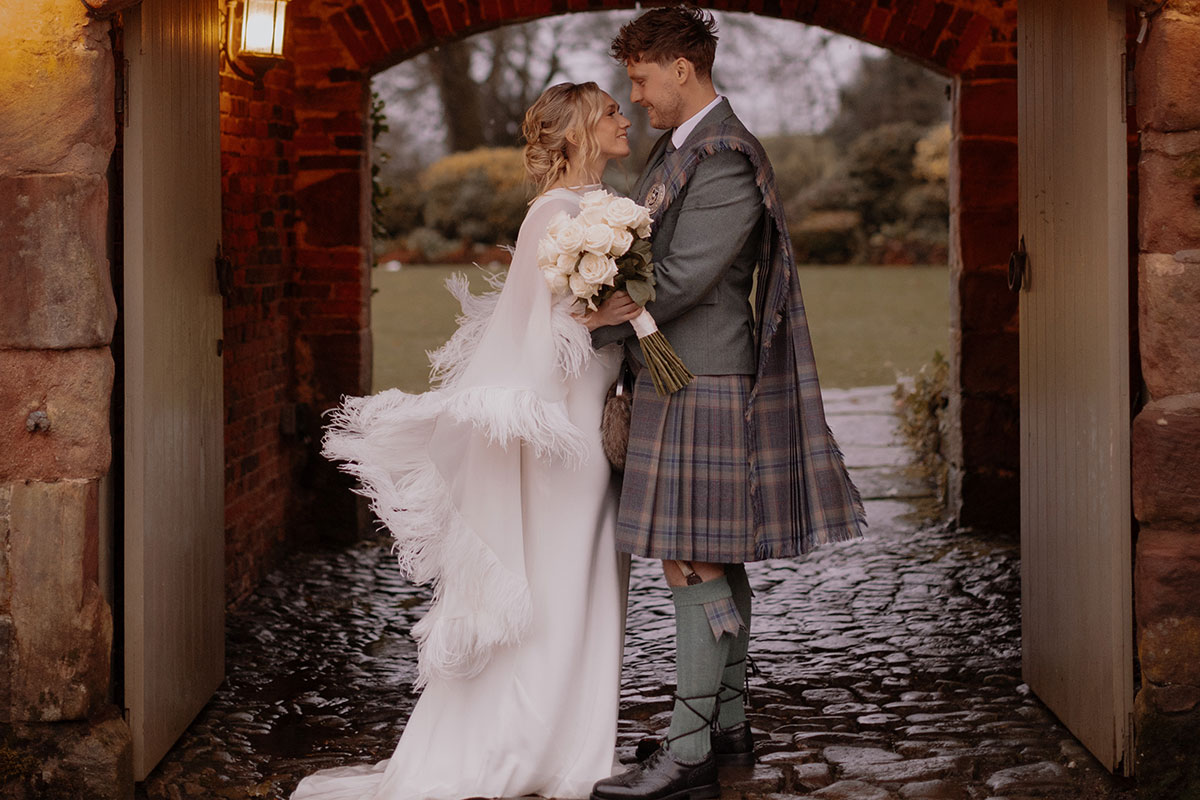 blonde bride in feathery cape and white wedding dress holds bouquet of white roses and stands smiling and facing groom in green jacket and red, blue, green kilt with matching fly plaid, both stood under outdoor stone archway