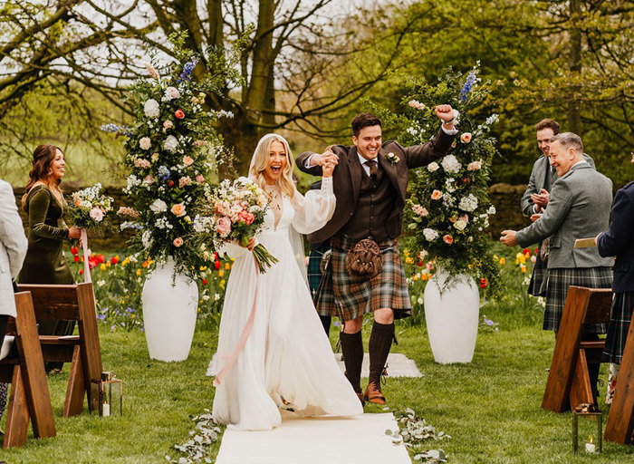 a bride and groom skip joyfully up an white carpet aisle in a flower filled garden setting