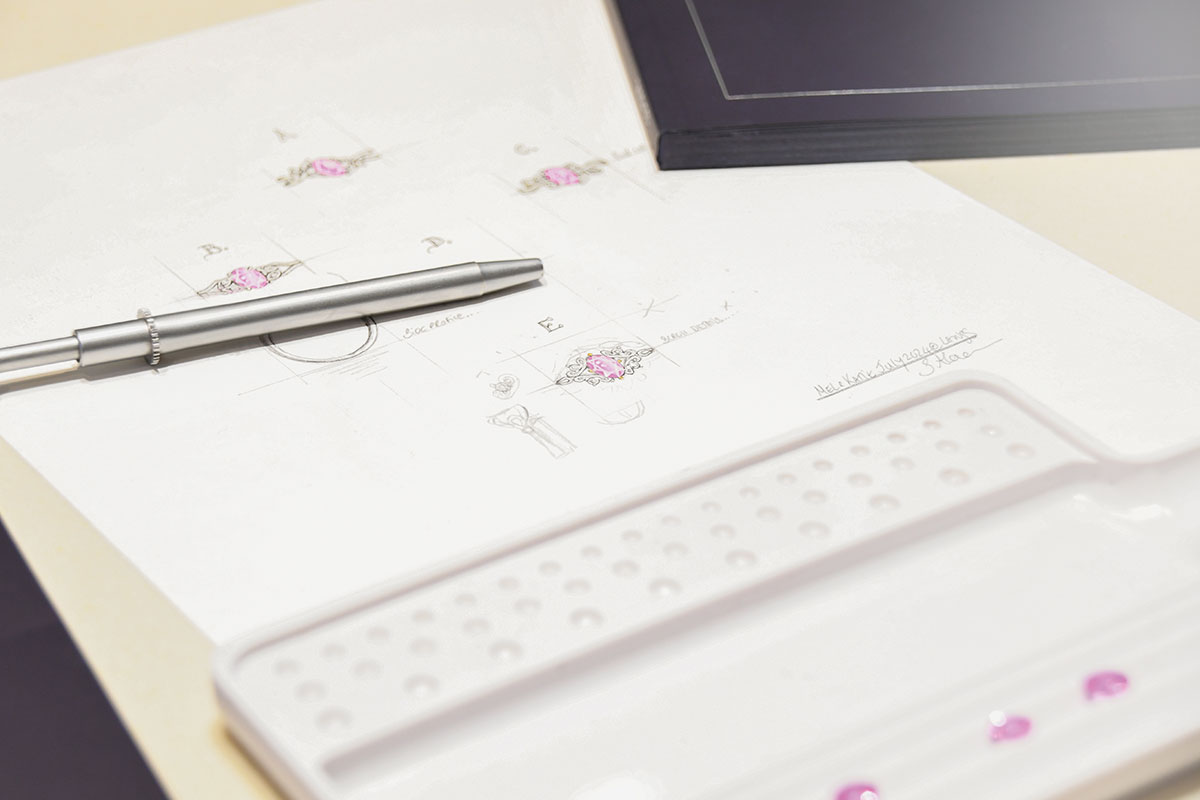 Bespoke engagement ring design sketches showing a pink gemstone ring alongside a jeweller's pen and loose stones