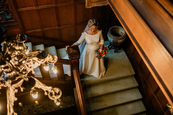 overhead shot of bride in white wedding dress and long veil holding pink bouquet as she walks down a carpeted staircase