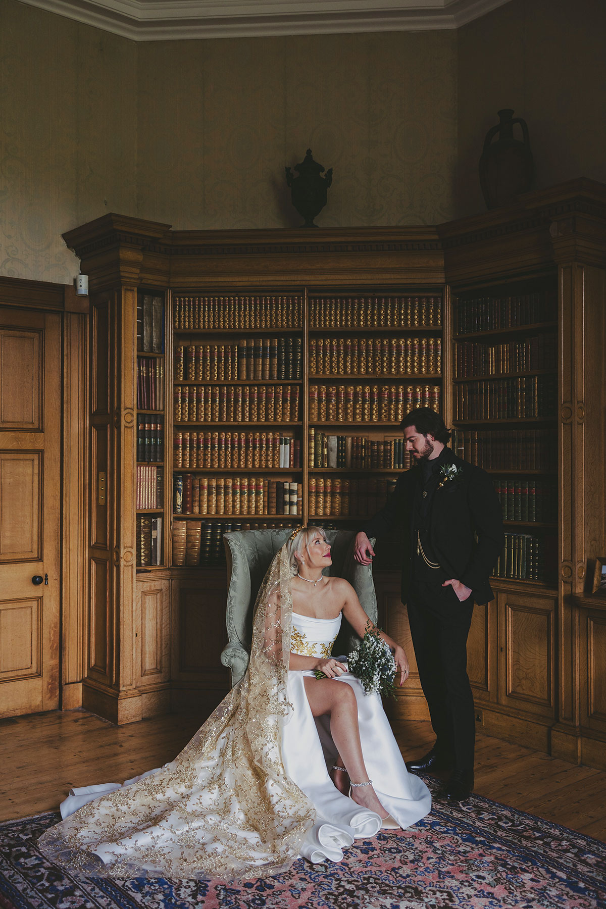 Bride seated in an ornate library in a gold-embroidered veil while the groom stands beside her, creating an elegant and dramatic wedding portrait.