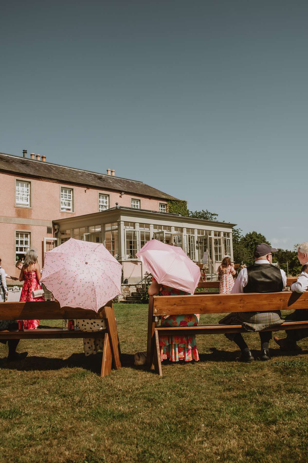 Wedding guests seated on wooden benches outside Ballogie House with the conservatory behind