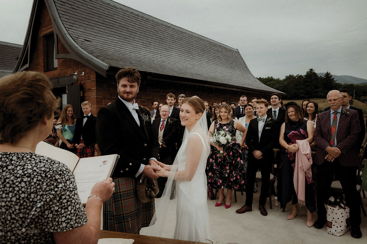 bride and groom holding hands in front of celebrant reading from book, formally dressed guests stood behind the couple looking on