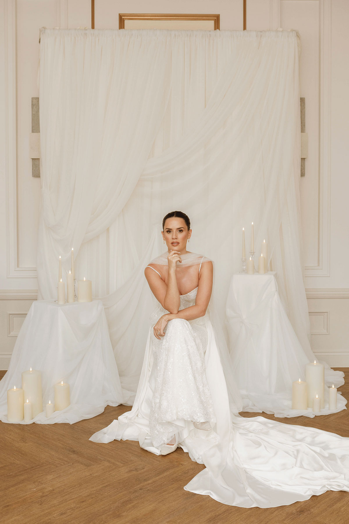Bride seated in an elegant white gown among draped white fabric and tall candles in a minimalist indoor wedding setup