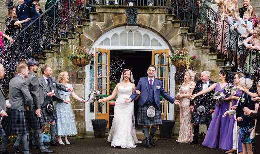 a bride and groom walking through a confetti shower below a stone staircase with wrought iron balustrade