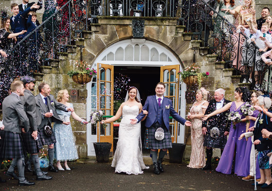 a bride and groom walking through a confetti shower below a stone staircase with wrought iron balustrade