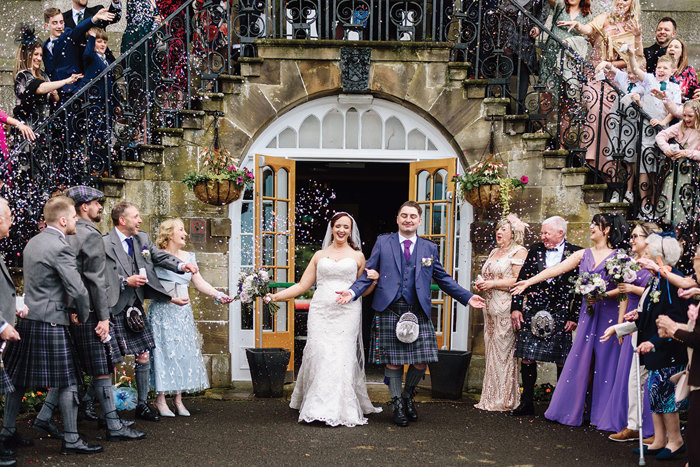 a bride and groom walking through a confetti shower below a stone staircase with wrought iron balustrade