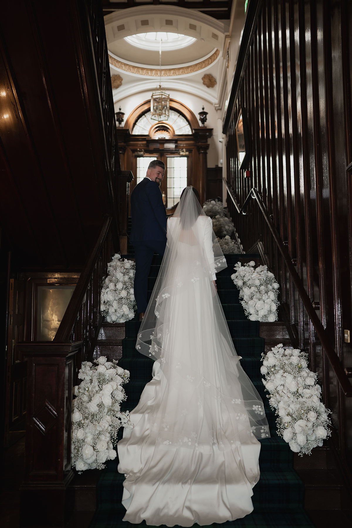 Bride and groom on the grand staircase at 1599 at the Royal College in Edinburgh, surrounded by white floral arrangements and elegant dark wood interiors