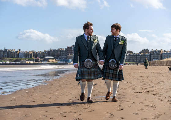 two grooms walking hand in hand along a sandy beach with St Andrews skyline in the background