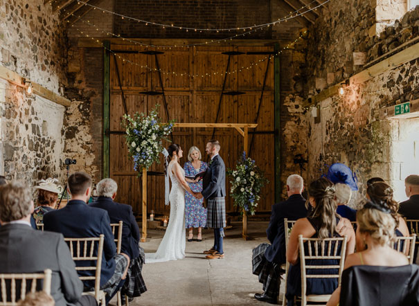 couple standing in front of a wooden arch in a barn during their wedding ceremony