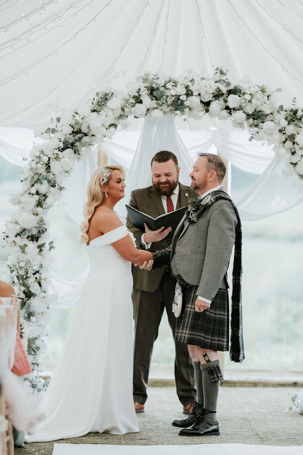 Couple stand with their celebrant beneath a white floral arch during the marquee ceremony.