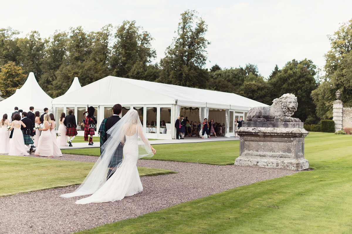 Bride, groom, wedding party and bag pipers walk across pebble path to get to white