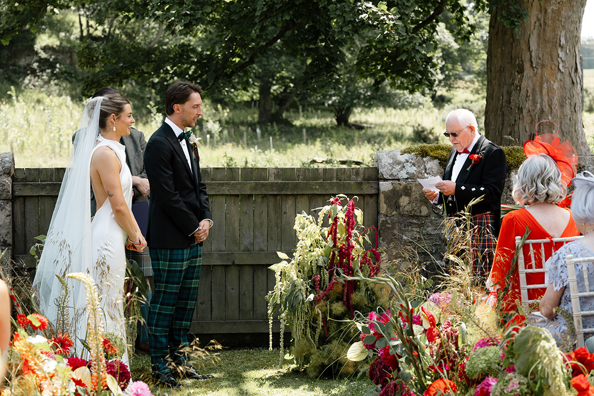 Grandfather does a reading during an outdoor orchard ceremony at Kinkell Byre, surrounded by vibrant red, pink and orange flowers at this colourful Fife barn wedding.