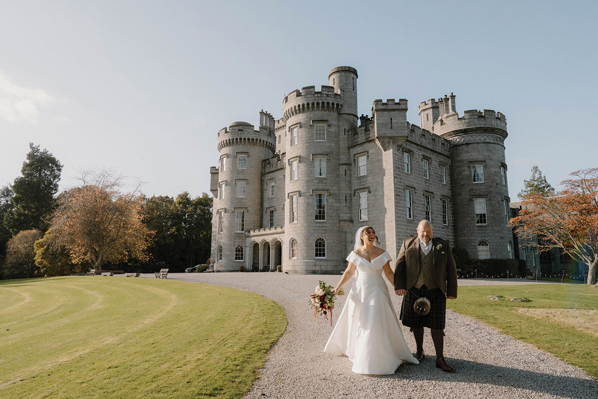 Bride and groom walking hand in hand outside Cluny Castle during an autumn Scottish castle wedding.