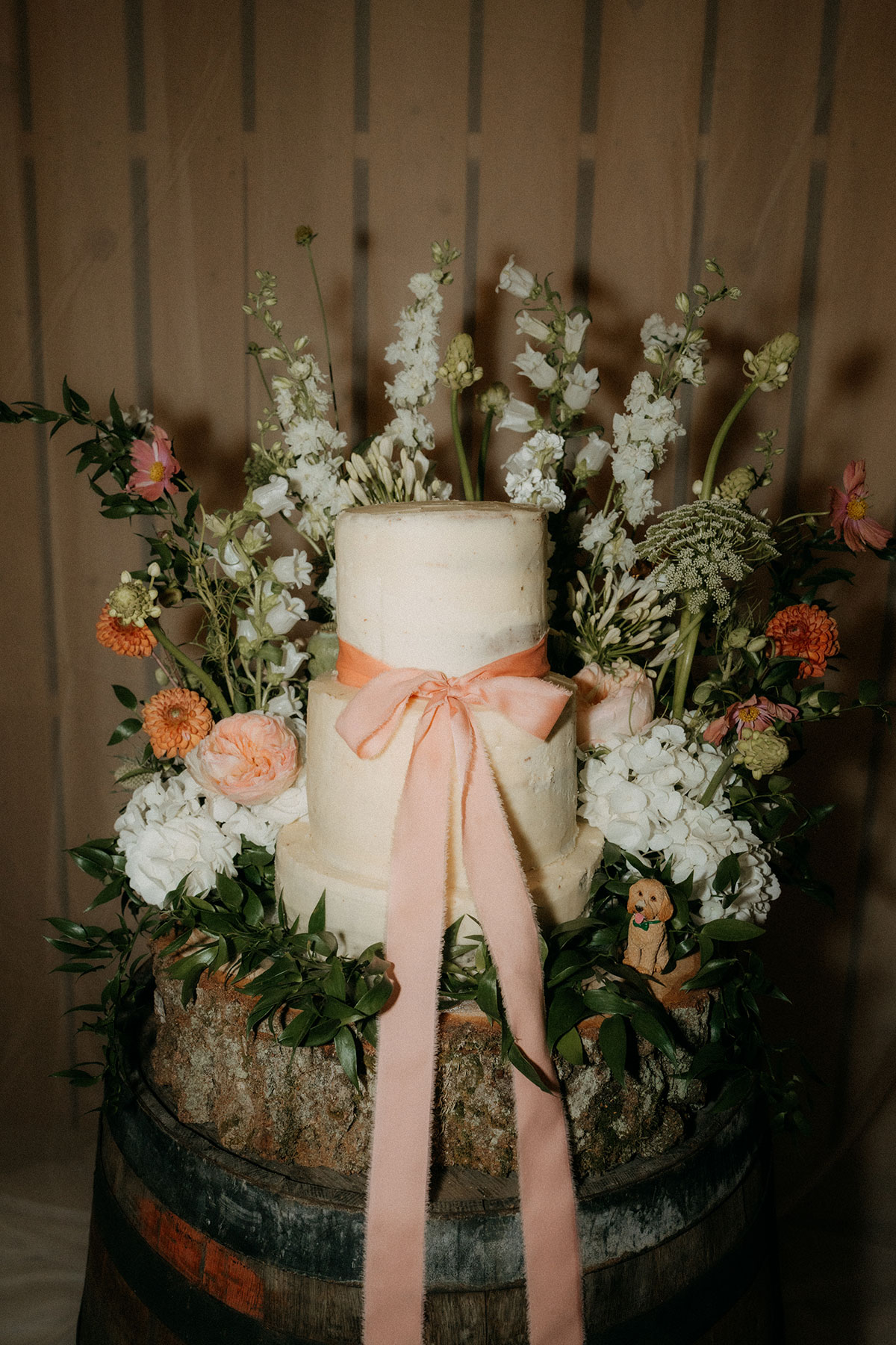 Three-tier wedding cake decorated with peach ribbon and fresh garden flowers on wooden barrel