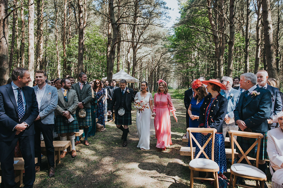 bride smiles as she walks down the aisle with her mum and dad as guests look on happily at harvest moon