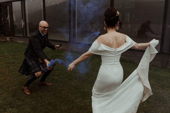 groom in blue, grey and white tartan kilt outfit crouches down on grass holding blue smoke stick as bride in white wedding dress dances in front