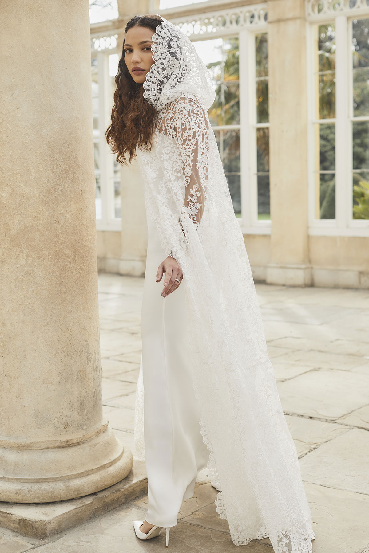 brunette curly haired woman stands in large stone tile room with large windows and looks back over her shoulder wearing silky column white gown with hooded lace cape