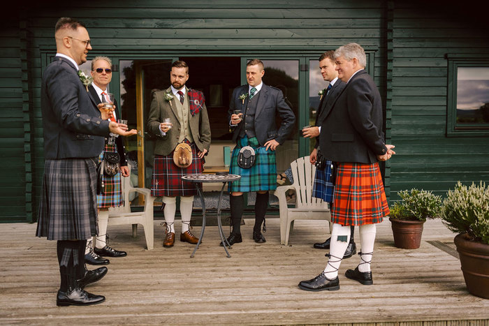 Men in kilts stand having a drink on decking in front of a green building 