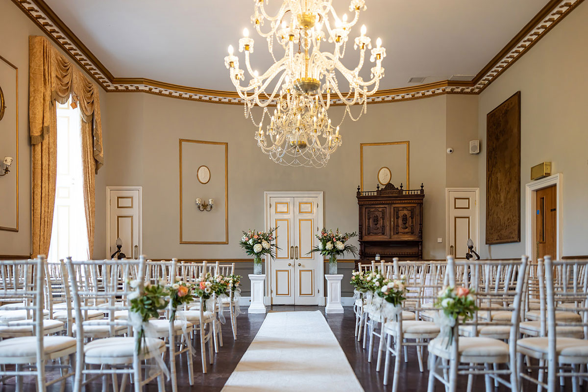 Ceremony setup inside Melville Castle featuring a crystal chandelier, white aisle runner, floral arrangements and Chiavari chairs in an elegant reception room