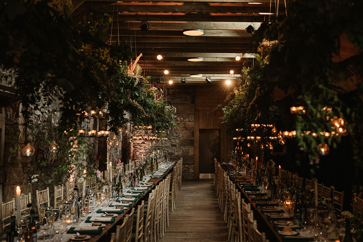 Two long banquet tables set beneath hanging greenery and candlelit glass baubles inside a rustic stone barn.