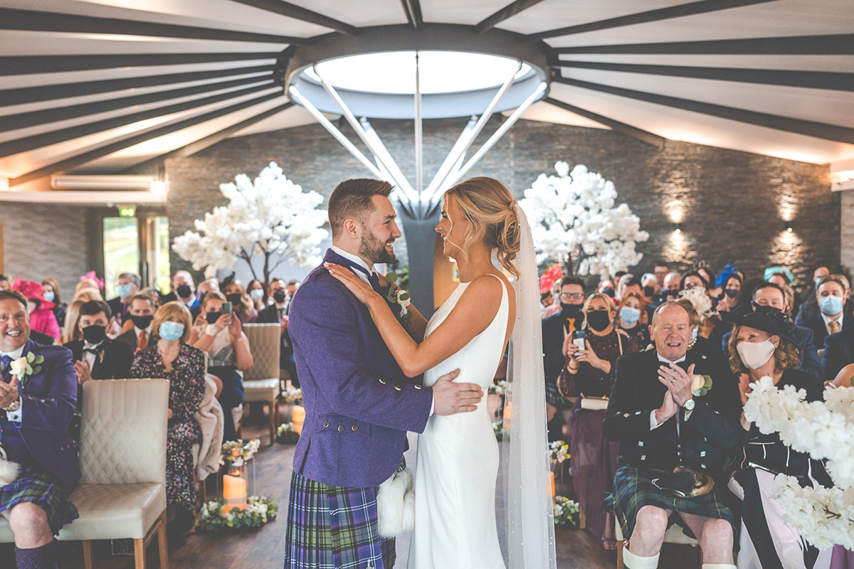 bride in sleek simple wedding dress and pearl veil stands with her hands on shoulders of groom in purple jacket and kilt outfit as they stand in front of friends and family seated in cream chairs behind them with white floral trees, one on either side of the couple