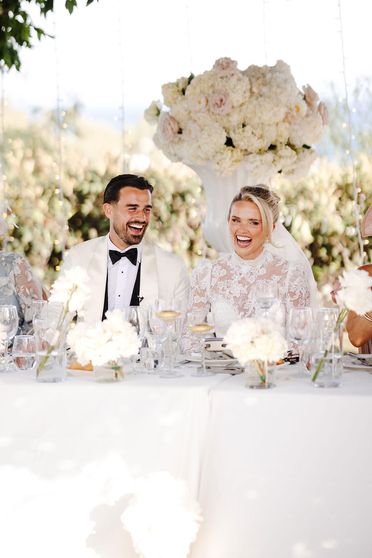 Bride and groom share a happy moment during speeches at a flower-filled outdoor reception