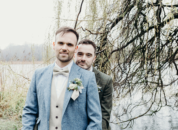 two grooms on their wedding day pose beside a body of water with overhanding branches and greenery