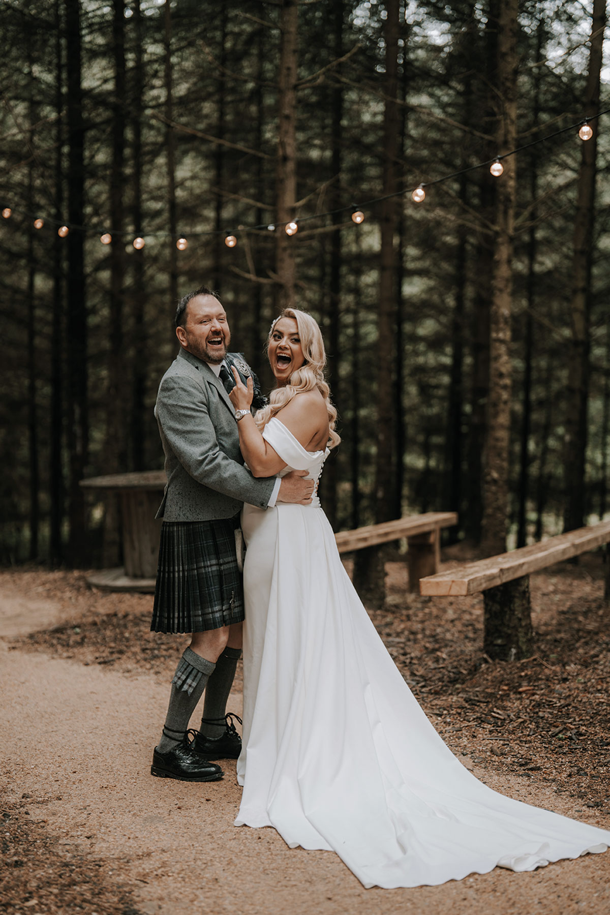 Bride and groom laughing together during their woodland photo session with string lights overhead.