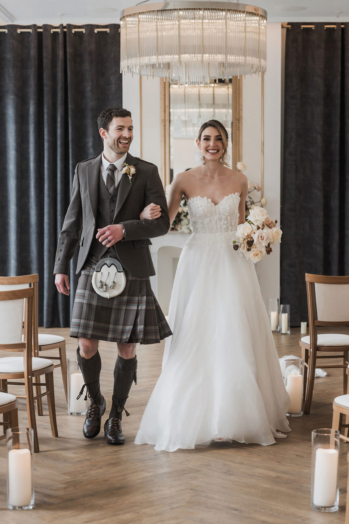 a bride and groom walking arm in arm up the aisle at the Exchange wedding venue in Glasgow. The brunette bride is wearing a wedding dress with floral applique on the bodice and the groom is wearing a brown tartan kilt outfit.