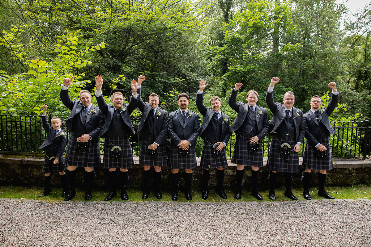 Groom and groomsmen in matching dark kilts and jackets cheer with raised arms outdoors, surrounded by green trees