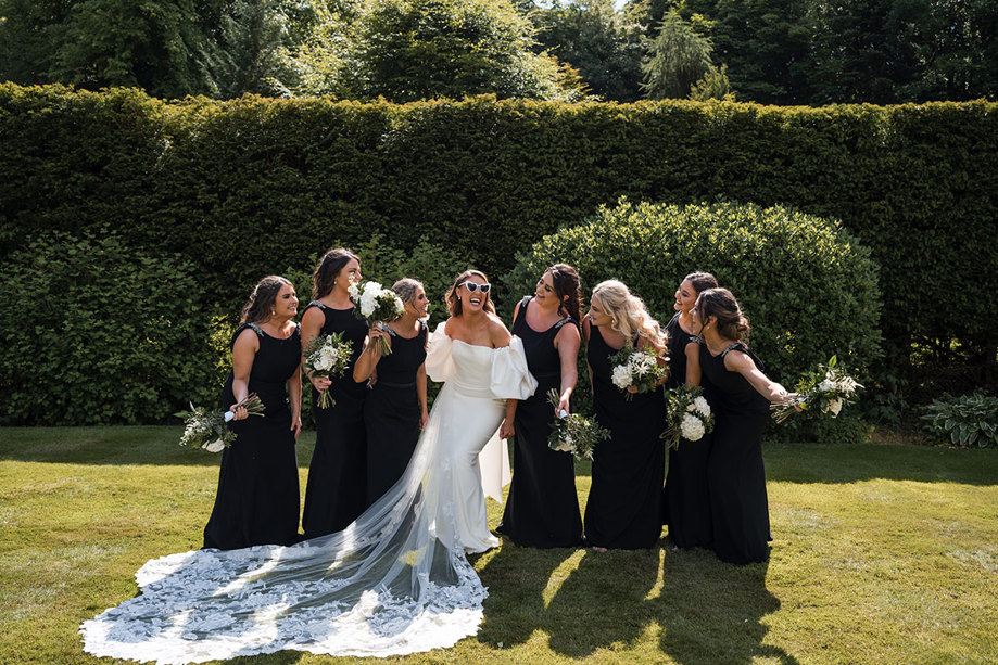 Bride wearing white love heart-shaped sunglasses laughs with her bridesmaids dressed in black and holding flowers