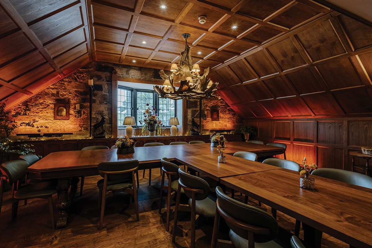 a dimly lit room with oak panelled walls and wooden dining tables formed into a T-shape with small flower arrangements placed at the centre points of each