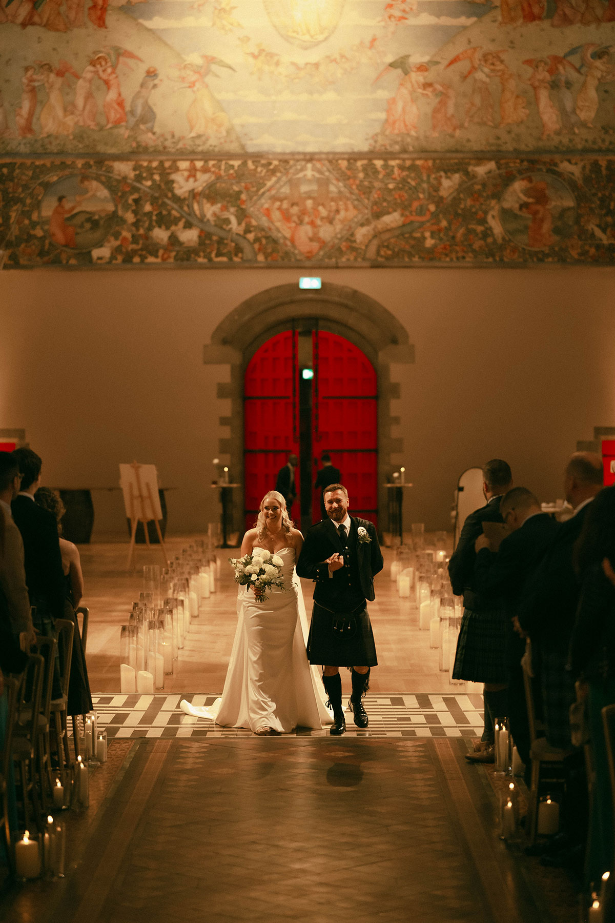 bride and groom walking down candlelit aisle in historic wedding venue with red doors