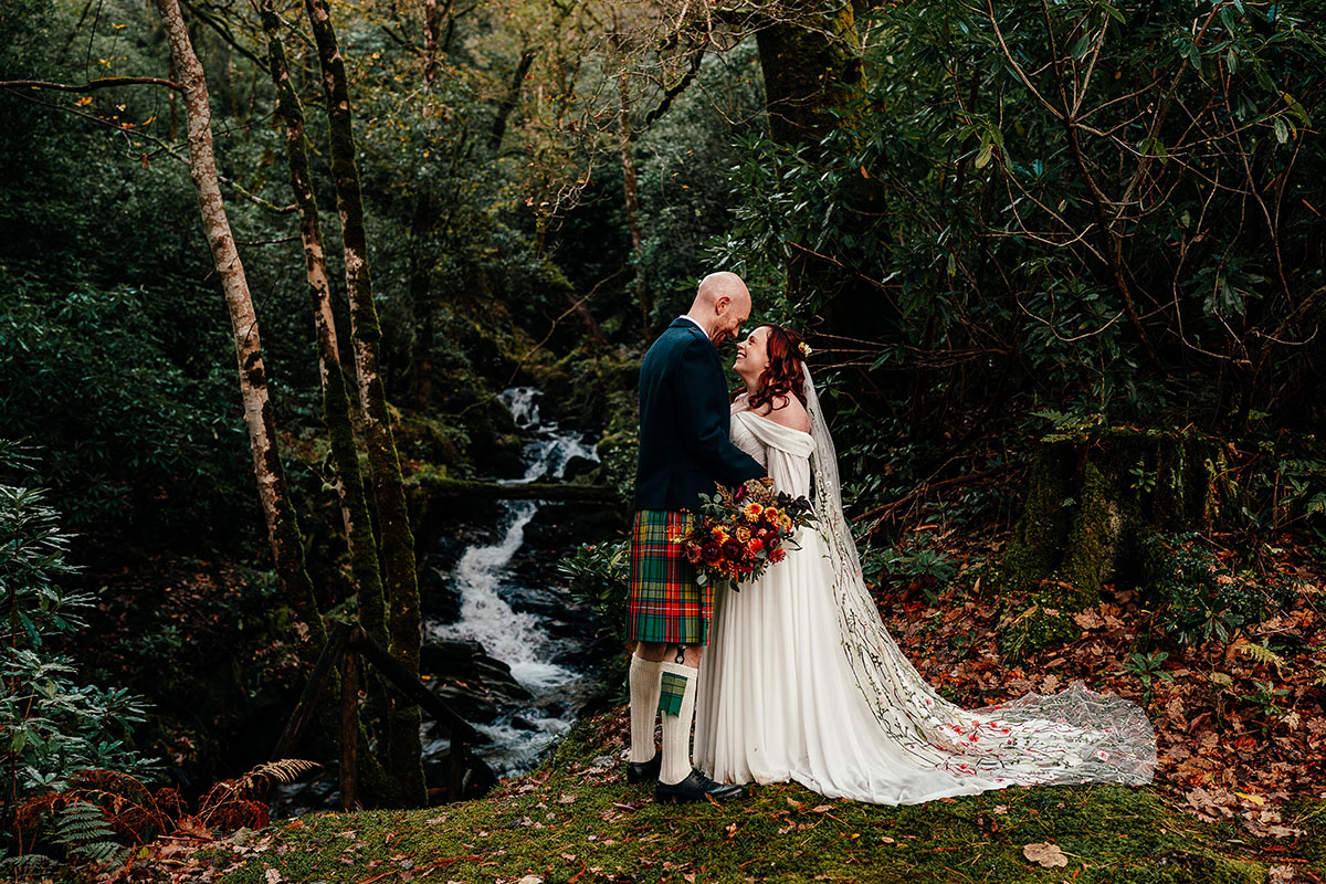Bride and groom embrace beside a woodland waterfall, surrounded by lush greenery and autumnal florals
